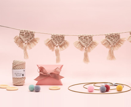 Beige Makramee-Girlande, Dekoringe und bunte Garn-Pompons. Hängende Makramee-Girlande mit Garn, eine Geschenkschachtel, verschiedene Dekoringe und Verpackung mit Garn-Pompons. Fotosetting rosa Wand.