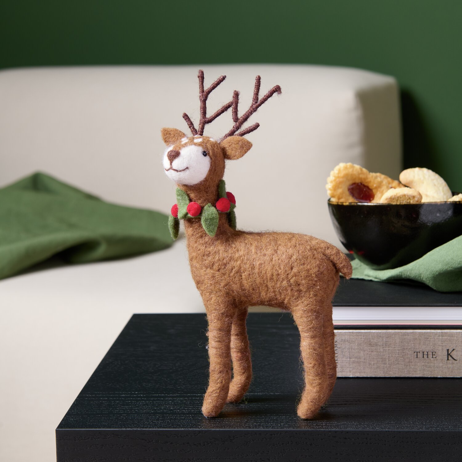 Filz-Hirsch mit Ilexkranz Braun: Dekorativer Hirsch aus Filz, stehend auf einem Tisch, mit grünem Ilexkranz.