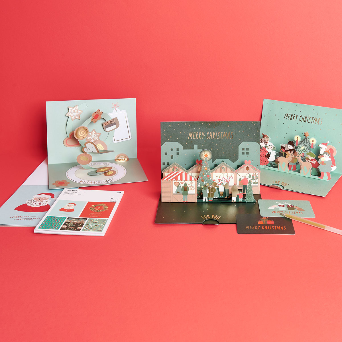 Drei unterschiedliche und weihnachtliche Pop-Up-Karten, dazu passendes Motivpapier und ein Gelstift. Fotosetting roter Hintergrund.
