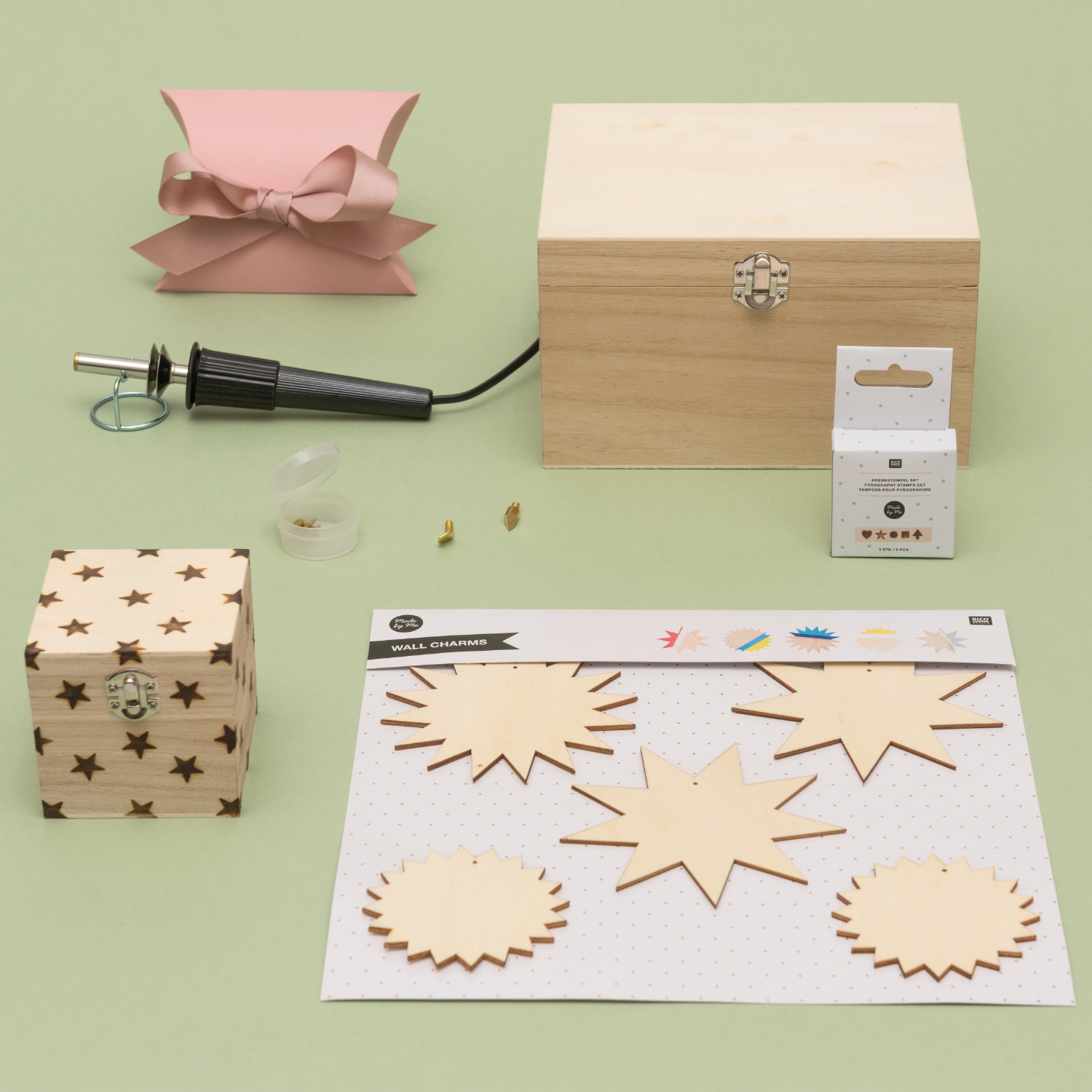 Ein Brandmalkolben mit Holzanhängern, einer großen Holzkiste und einer kleinen Holzkiste mit kleinen Sternen, sowie eine pinke Geschenkschachtel. Fotosetting grüne Wand.