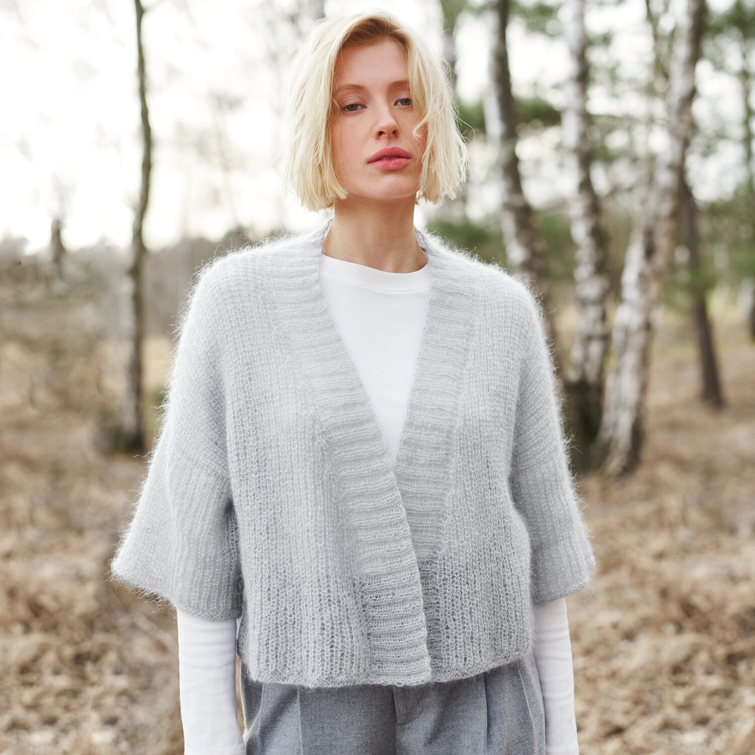 Strickset Kimono-Jacke Modell 19 aus Lovewool No. 21 von Rico Design Strickset Kimono-Jacke Modell 19 aus Lovewool No. 21: Graue, kurzärmelige Jacke im Wald getragen, kombiniert mit weißem Shirt und grauer Hose.