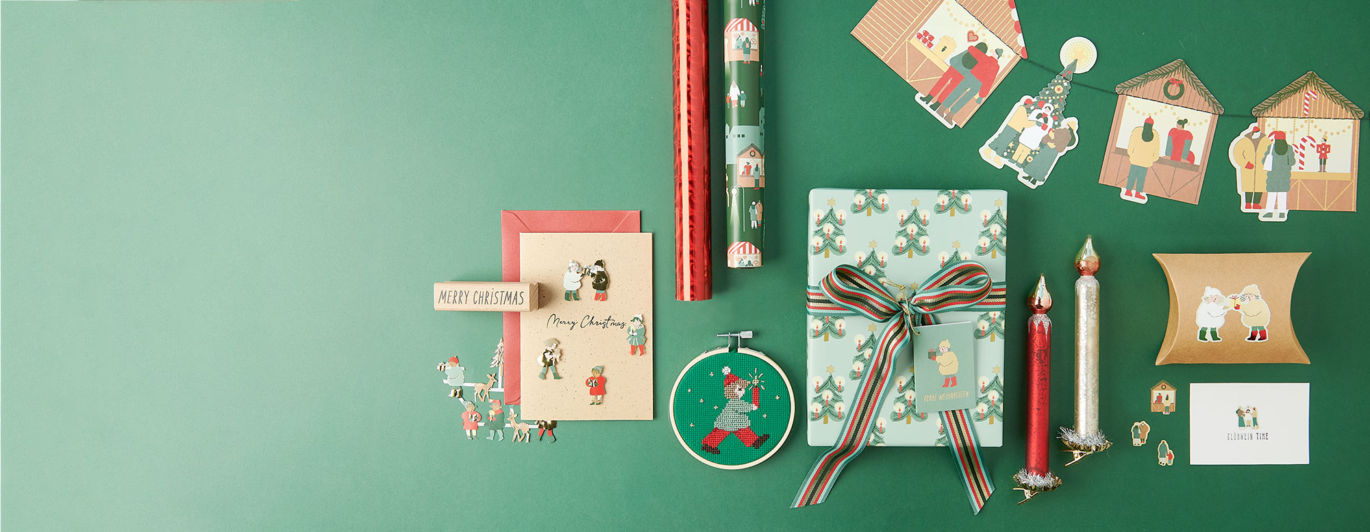 Verschiedene weihnachtliche Artikel zum Dekorieren, Basteln und Verschenken. Ein verpacktes Geschenk, Geschenkpapierrollen, eine verzierte Weihnachtskarte, ein Stickring, eine verzierte Geschenkschachtel, Kerzen und eine Girlande alle in weihnachtlichem Stil.