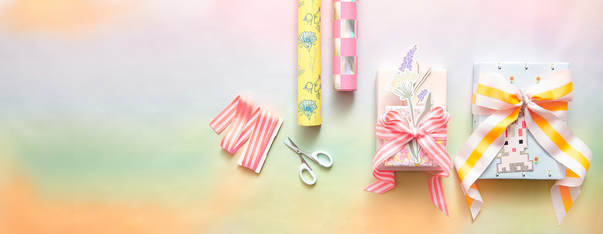 Zwei verpackte Geschenke mit Geschenkanhänger und Schleife, daneben zwei Geschenkpapierrollen, Geschenkband und eine Schere. Flatlay von Produkten.