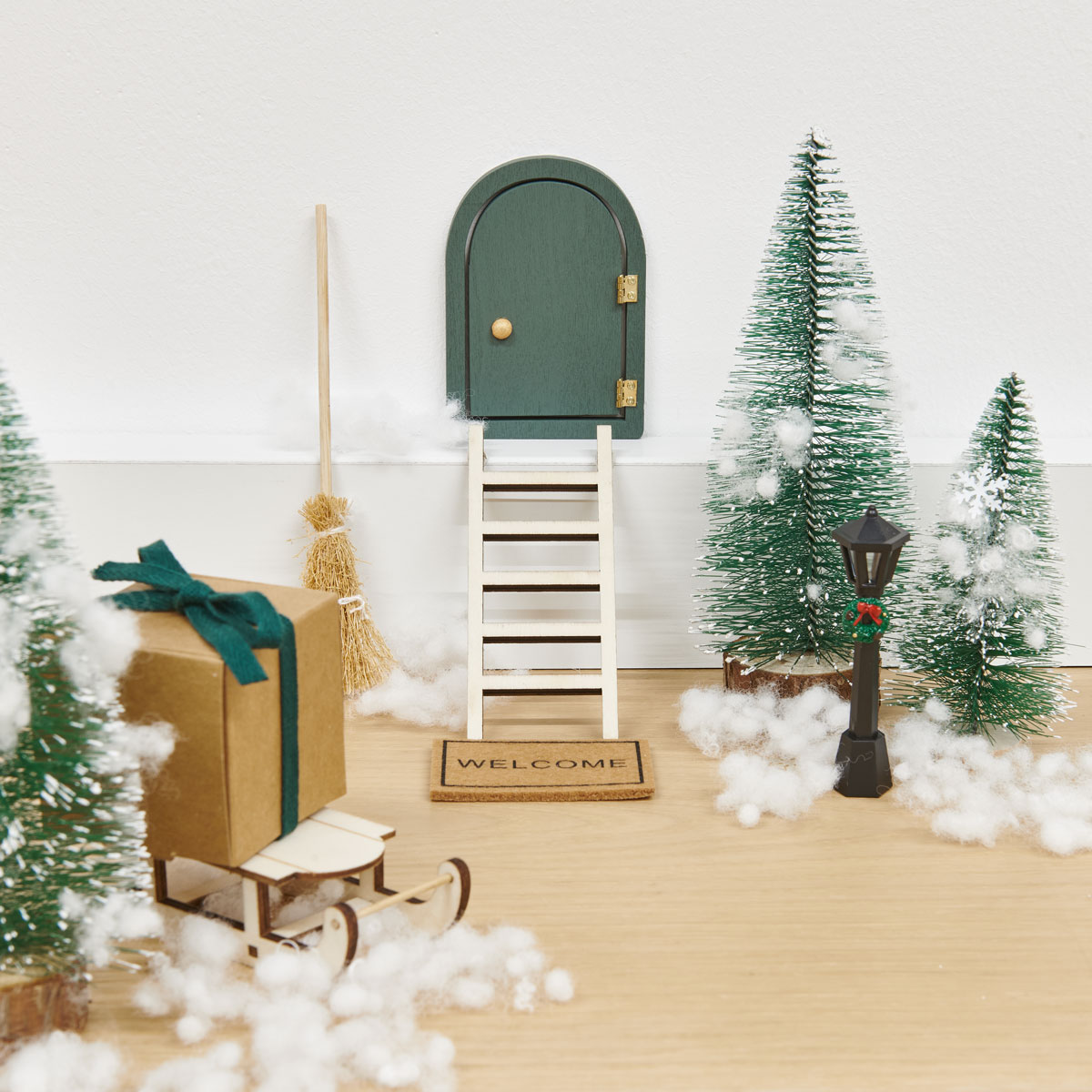 Ein Miniatur-Wichtel-Hauseingang bestehend aus einer Tür, Treppe, Tannenbäumen, Schlitten, Geschenk und Watte. Fotosetting Fußleiste.