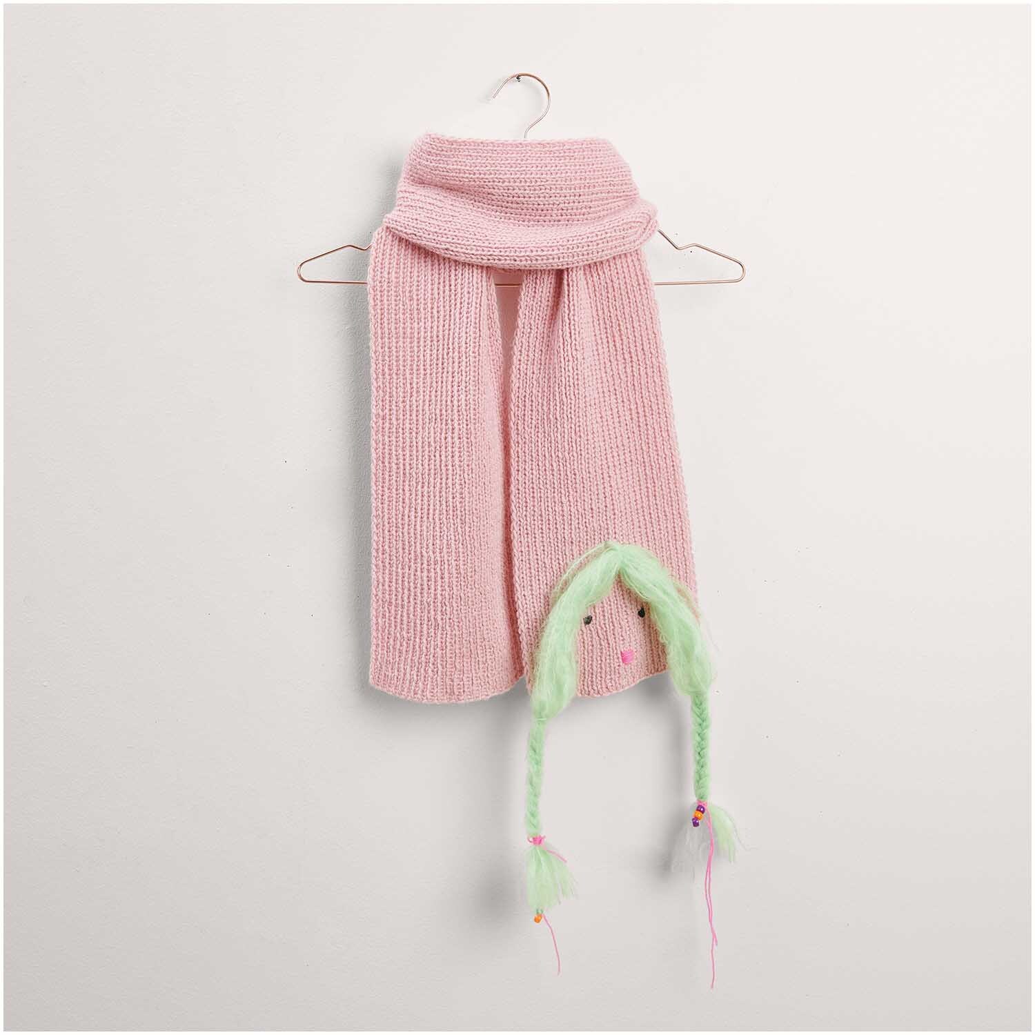 Ein rosa Strickschal von "Made by Me", Modell 29 aus der Kollektion Nr. 17, hängt an einem Kleiderbügel. Der Schal hat ein verspieltes Design mit grünen Zöpfen am Ende.