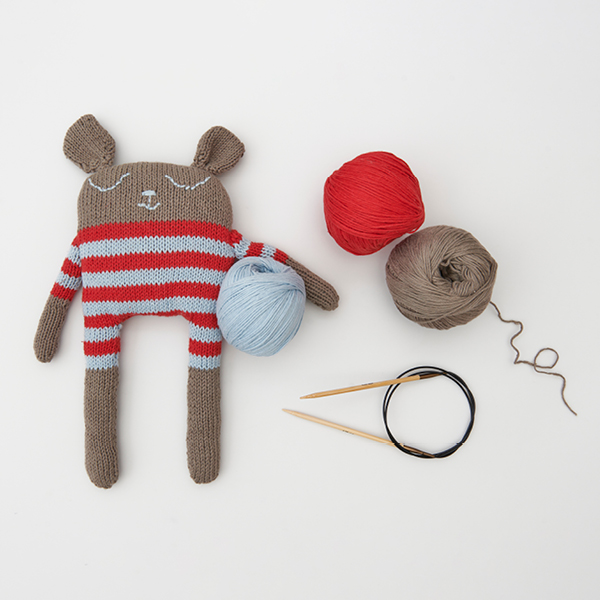 Ein gestricktes Kuscheltier, daneben drei dazugehörige Garnknäuel und eine Rundstricknadel. Flatlay von Produkten.