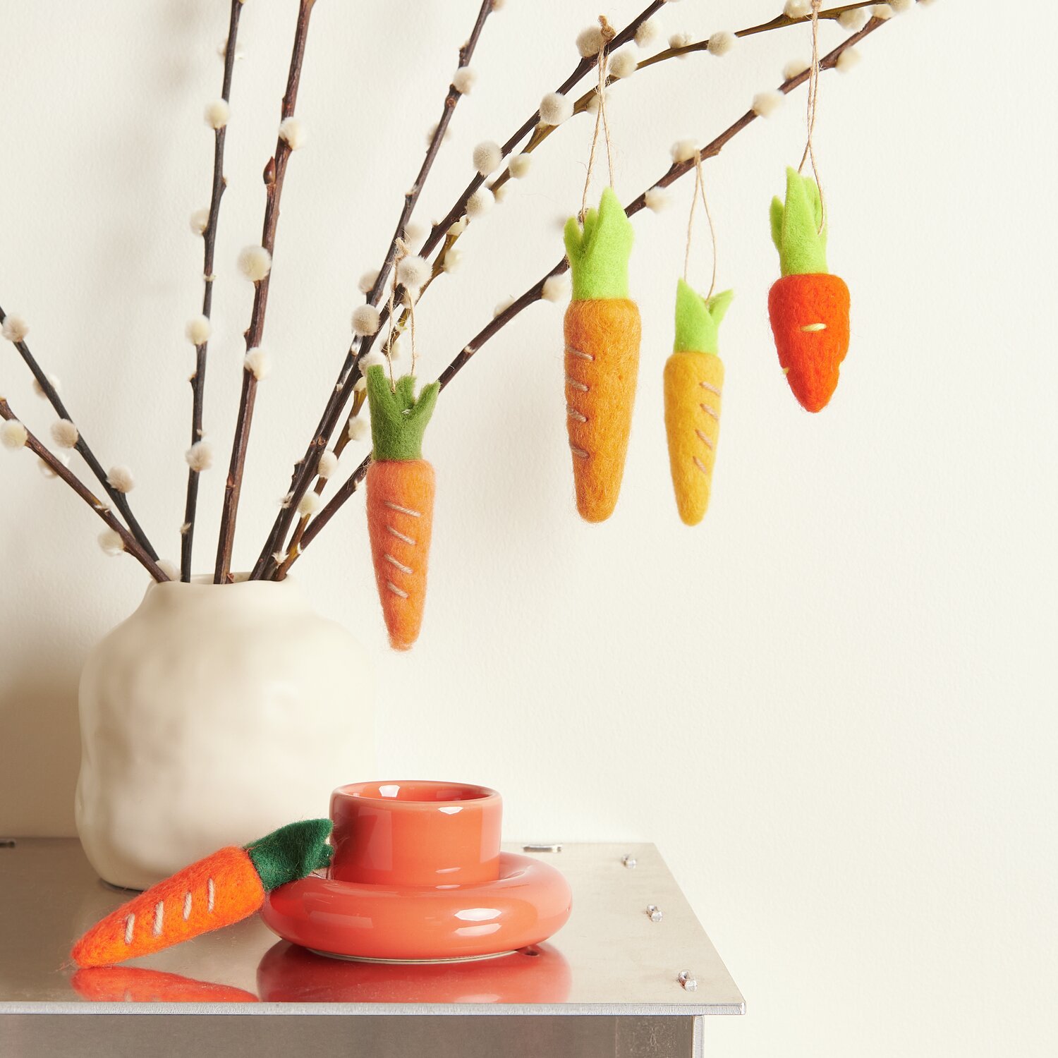 Aufhänger Möhre, 5-teilig: Filzkarotten in Orange und Grün hängen an Zweigen neben einer Vase.