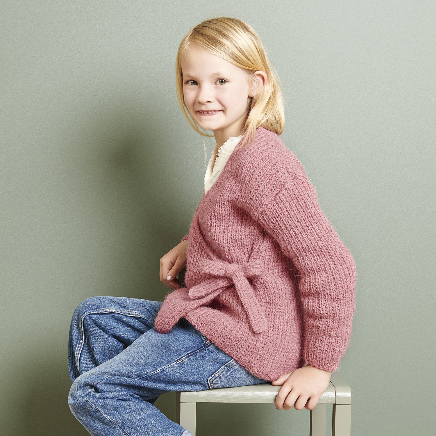 Strickanleitung Wickeljacke aus Soft Alpaca: Mädchen in rosa Wickeljacke, sitzt auf einem Stuhl vor grünem Hintergrund.