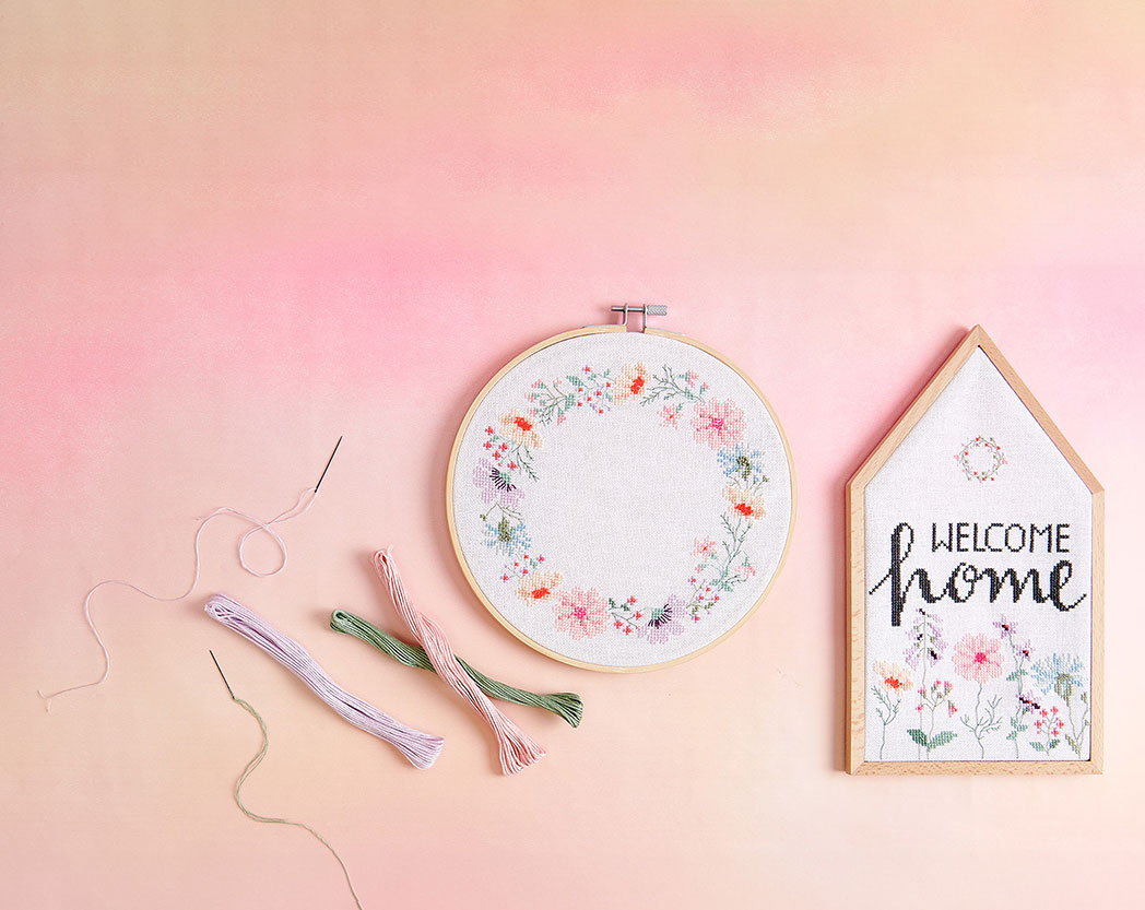 Ein Stickring mit einem Blumenkranz und ein Holzrahmen in Hausform mit Blumen und Schriftzug bestickt. Flatlay von Produkten.