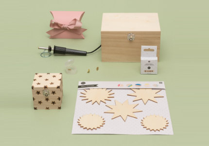 Ein Brandmalkolben mit Holzanhängern, einer großen Holzkiste und einer kleinen Holzkiste mit kleinen Sternen, sowie eine pinke Geschenkschachtel. Fotosetting grüne Wand.