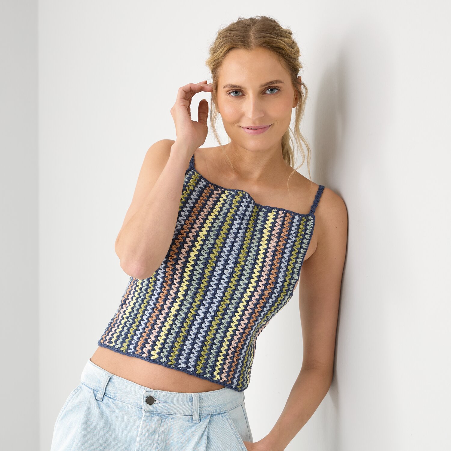 Häkelset Top Modell 03 A: Frau trägt bunt gestreiftes Top, lehnt lächelnd an weißer Wand.