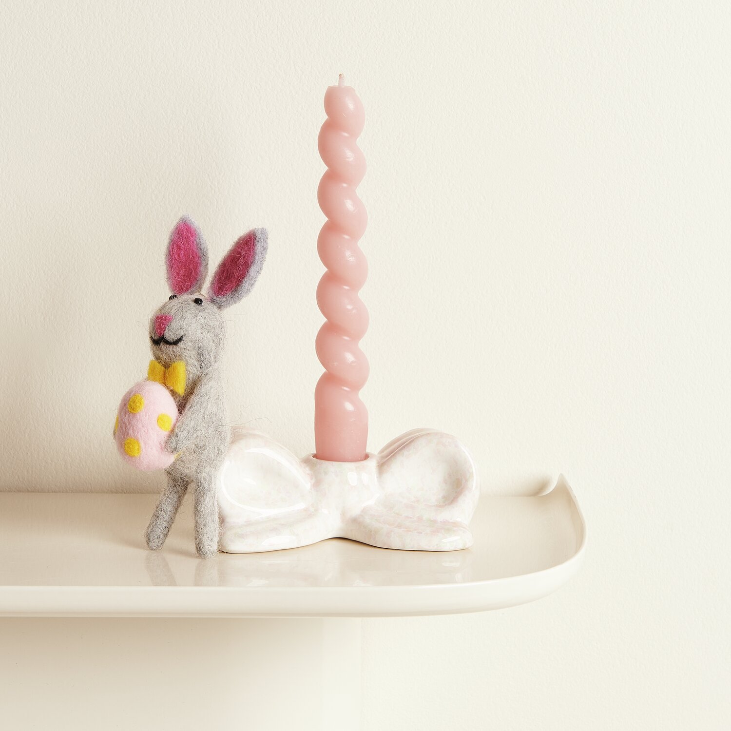 Aufhänger Filz-Hase mit Osterei und gelber Fliege neben rosa Kerze auf weißem Regal.