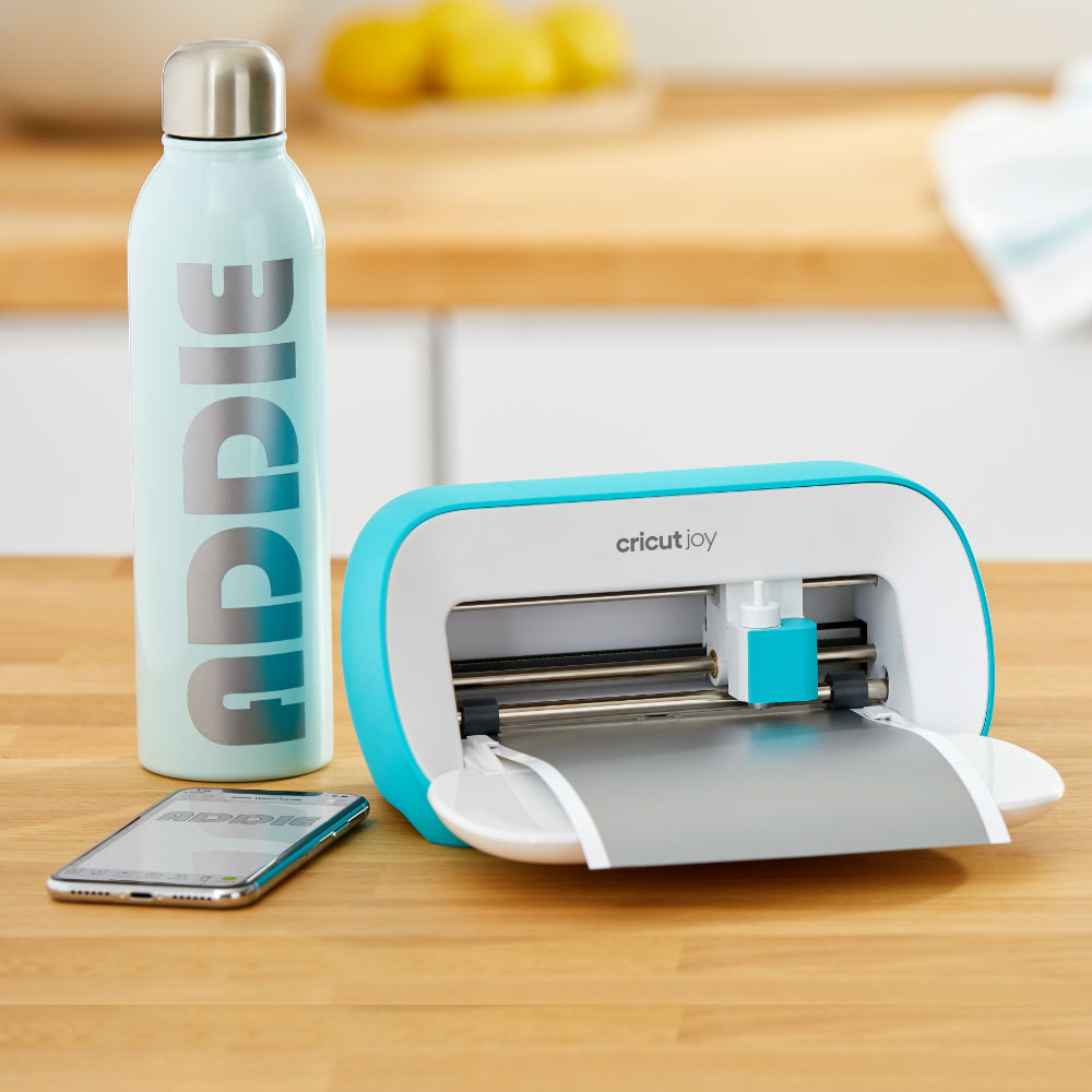 Ein kleiner Schneideplotter schneidet graues Papier, daneben steht eine Flasche und ein Handy.