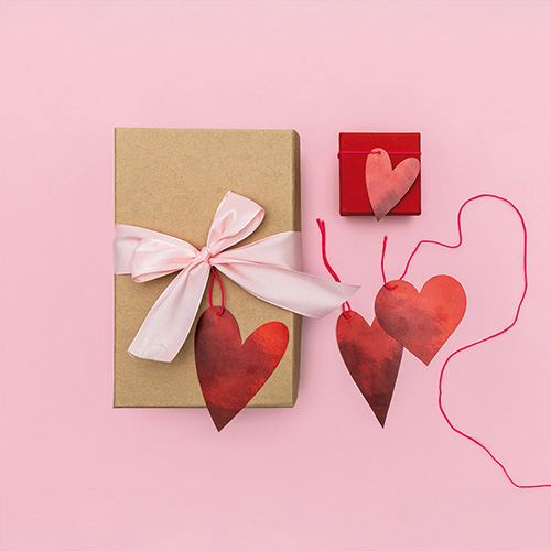Verpacktes Geschenk mit rosa Schleife und Herz-Anhänger und kleines verpacktes Geschenk mit kleinem Herz-Anhänger. Flatlay von Produkten.