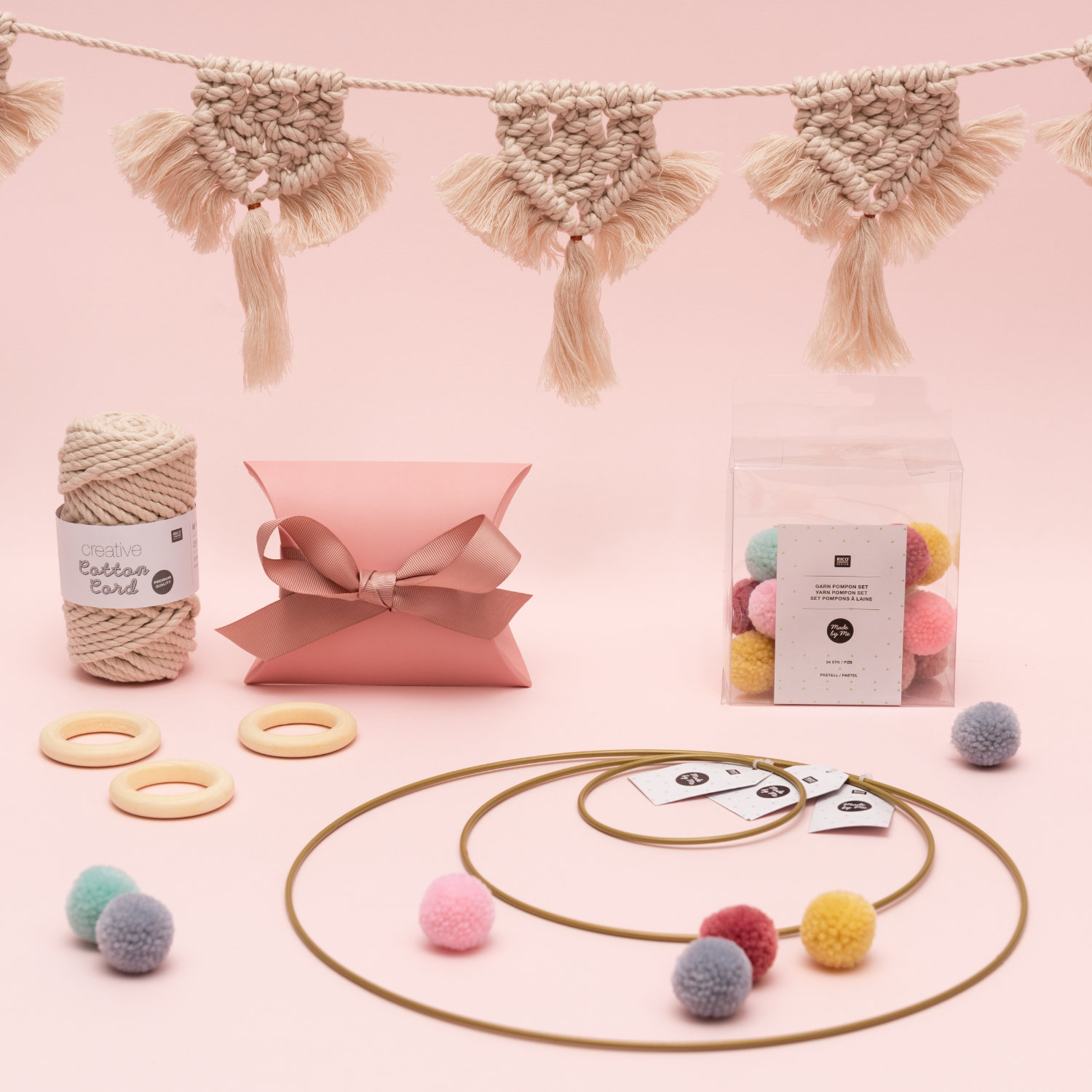 Hängende Makramee-Girlande mit Garn, eine Geschenkschachtel, verschiedene Dekoringe und Verpackung mit Garn-Pompons. Fotosetting rosa Wand.