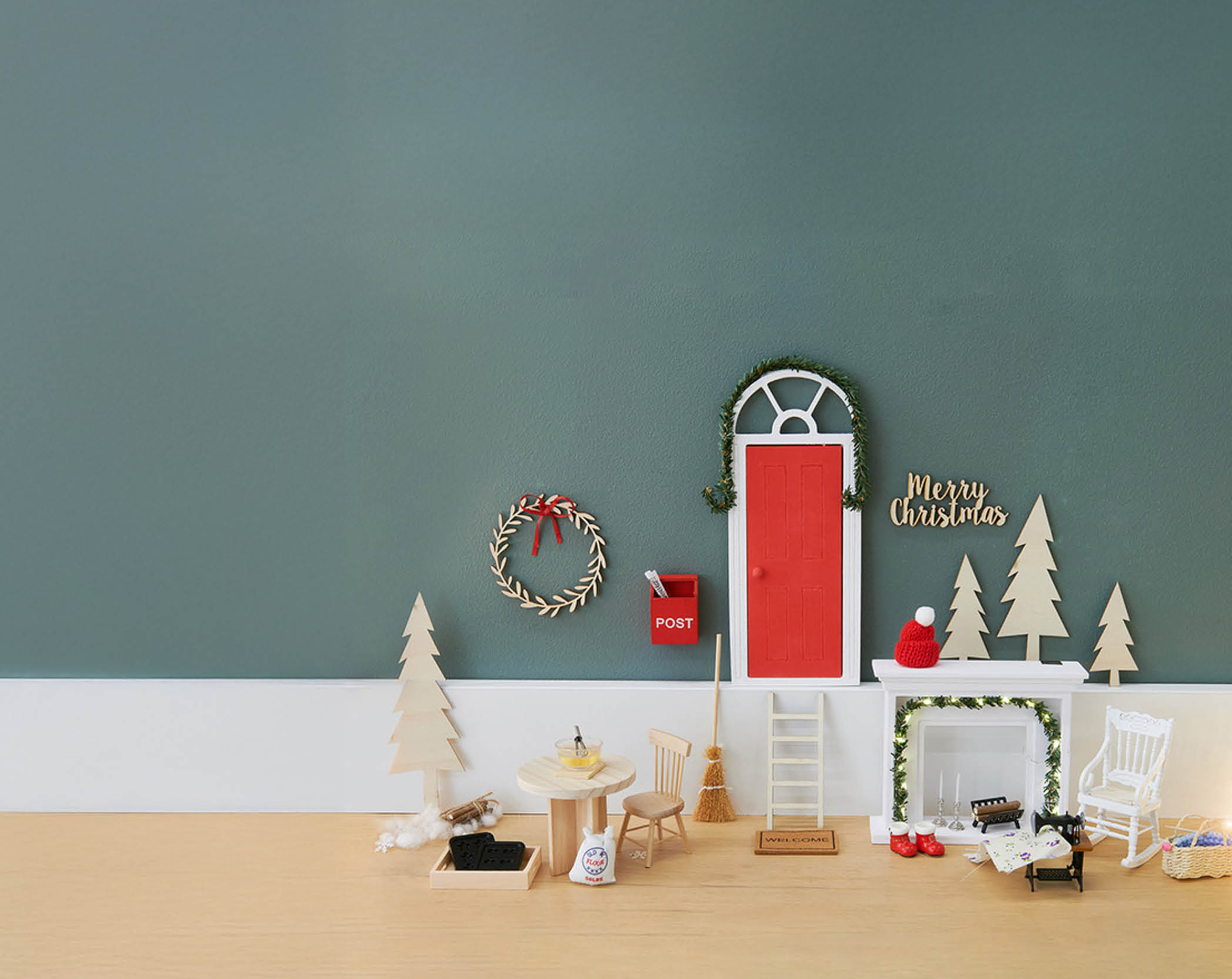 Ein Wichtel-Hauseingang mit einer roten Eingangstür, einem Briefkasten, einer Leiter, einem Kamin, sowie Tisch und Stuhl. Fotosetting Fußleiste und türkise Wand.