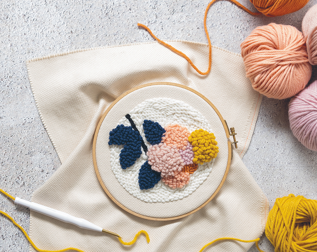 Ein Stickring mit Punch-Needle Technik bestickt, drumherum mehrere Garnknäuel und eine Punch-Needle. Flatlay von Produkten.