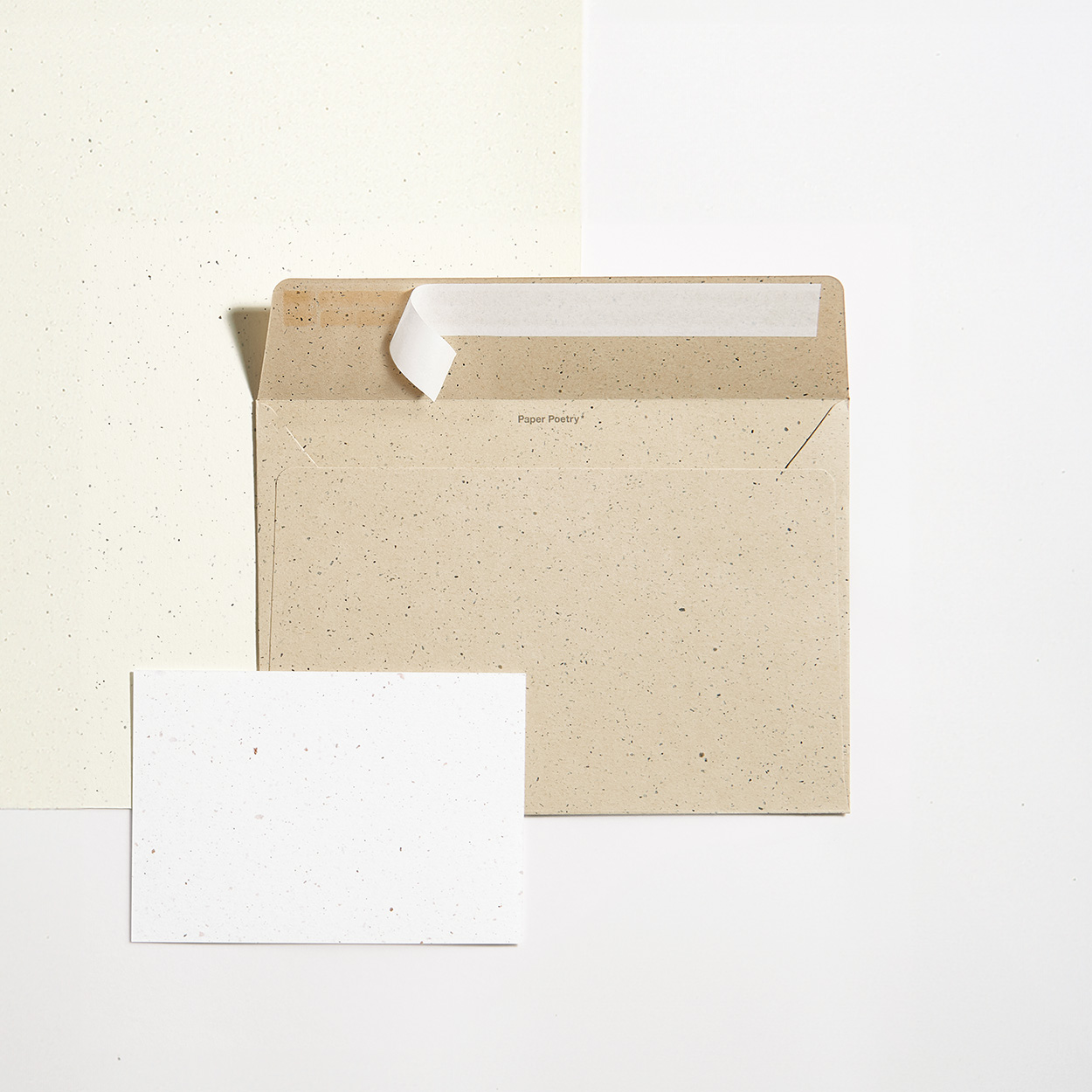 Ein bräunlicher Umschlag liegt auf creme-farbenem Papier und auf dem Umschlag liegt eine weiße Karte. Flatlay von produkten.