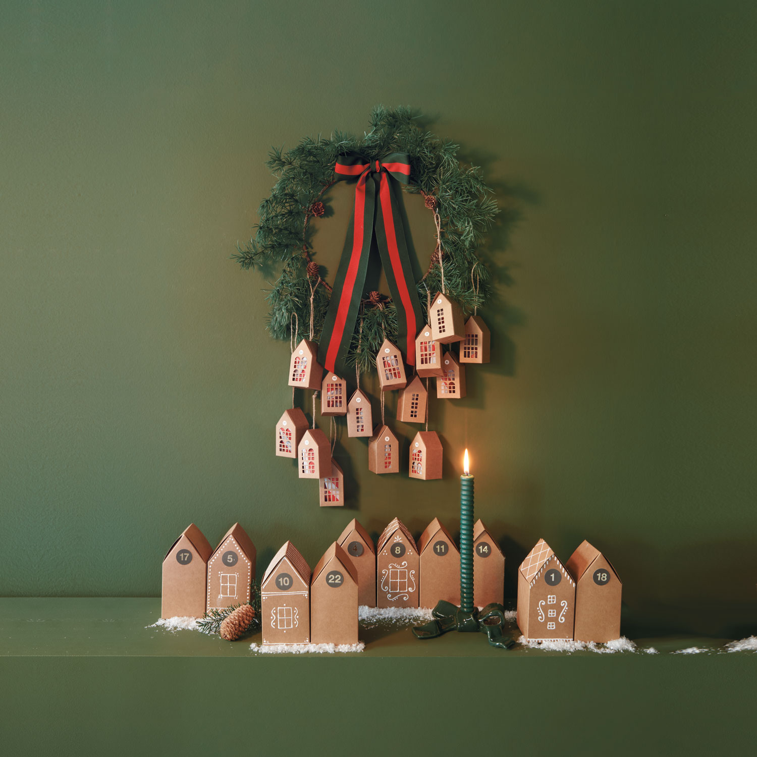 Ein Adventskalender bestehend aus kleinen und großen Pappschachteln in Hausform, sowohl stehend als auch an einem Kranz hängend. Fotosetting grüner Hintergrund.