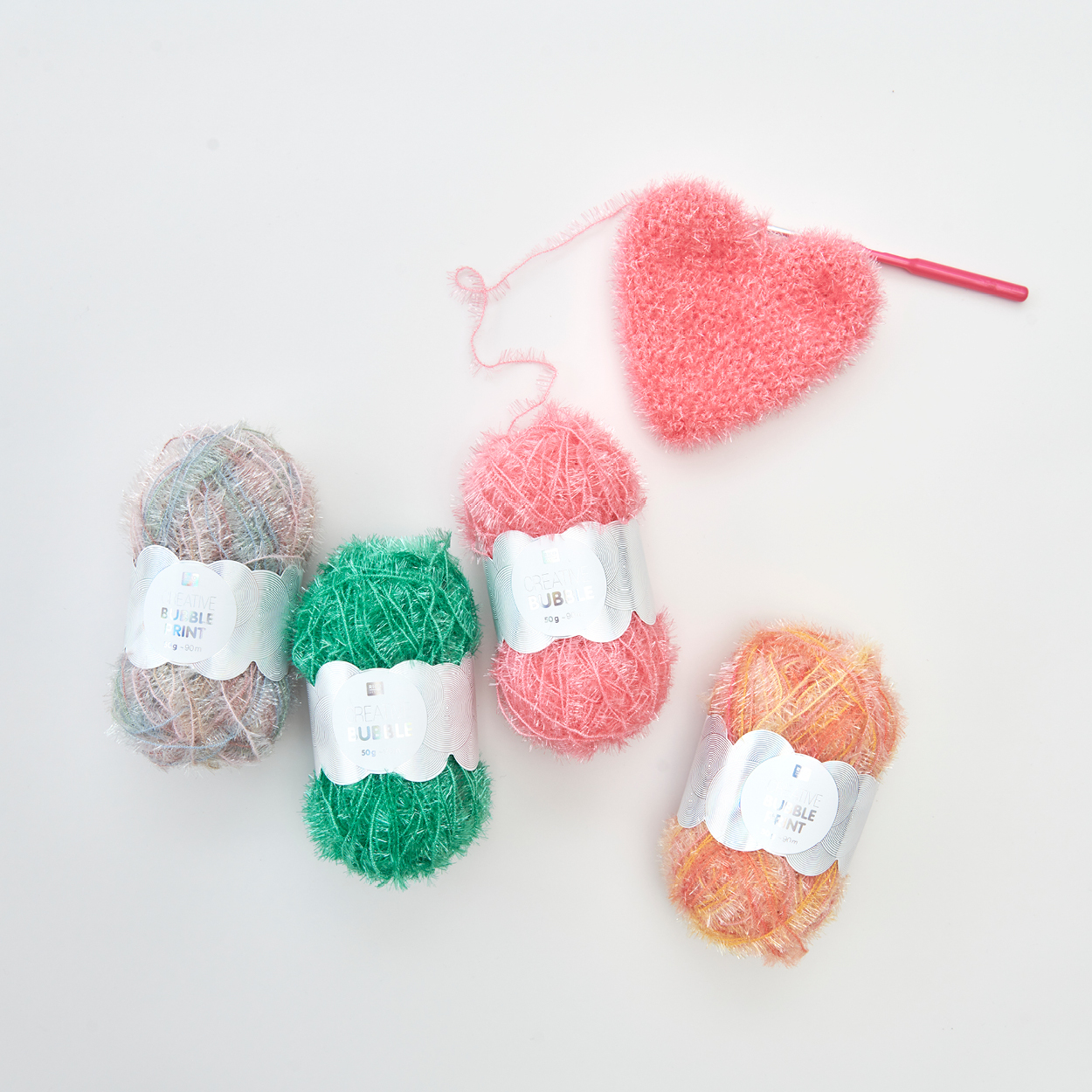 Vier anders farbige Schwammgarne mit einem pinken gehäkelten Herz mit Häkelnadel. Flatlay von Produkten.