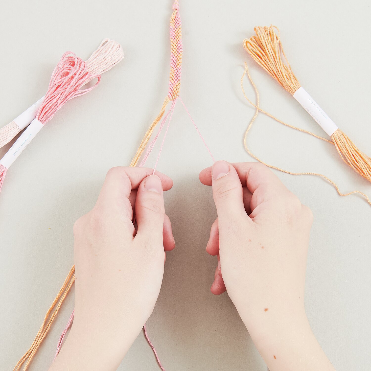 DIY Freundschaftsarmbänder Set Fashion Colours: Hände flechten bunte Fäden in Rosa und Orange.