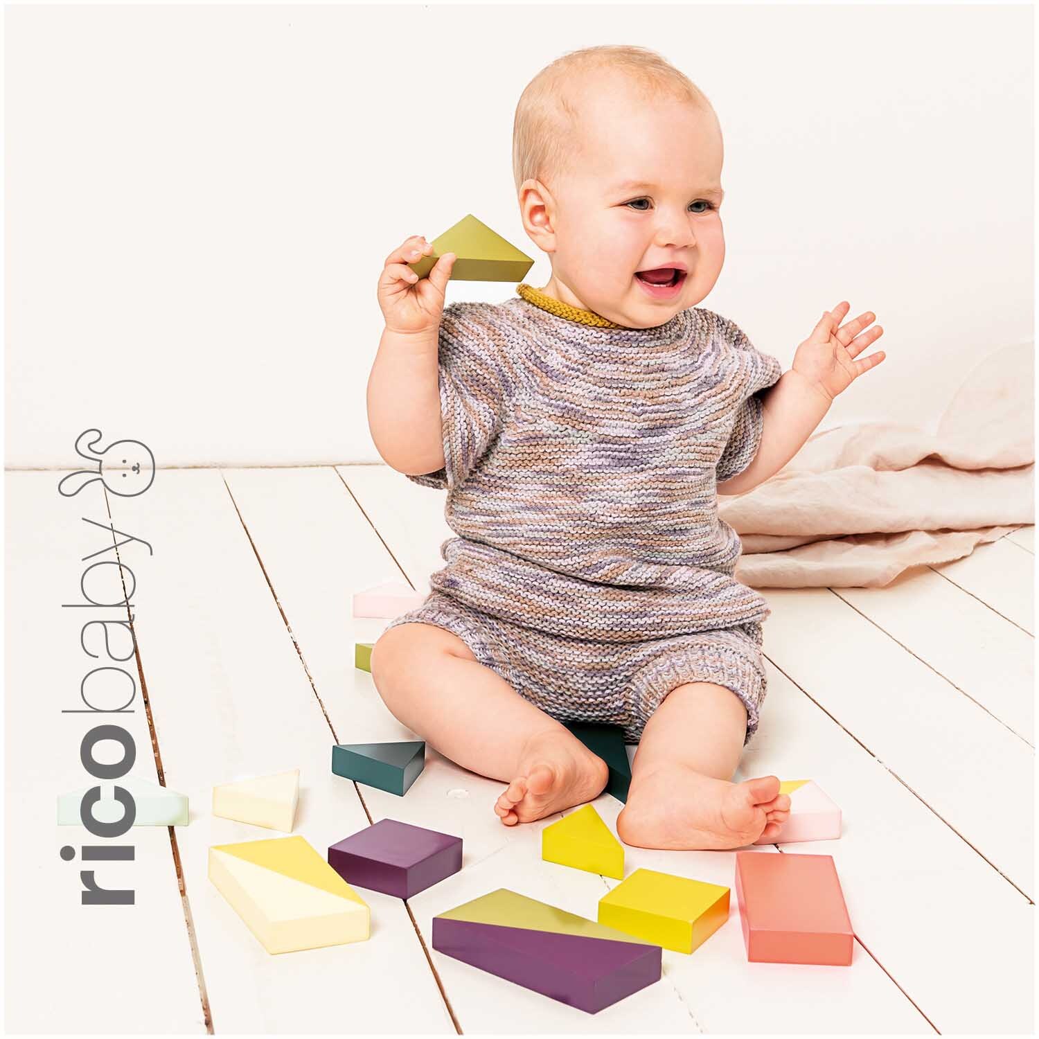 Rico Baby Cotton Soft Print dk: Ein Baby spielt auf Holzboden mit bunten Bauklötzen, trägt gestrickte Kleidung.
