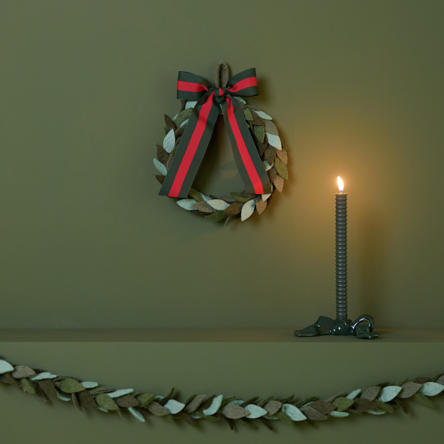 Filz-Blätterkranz Grün Ø25cm von Rico Design Filz-Blätterkranz Grün mit Keramik Kerzenhalter und Schleife von Rico Design, dekoriert auf einem Regal mit brennender Kerze.
