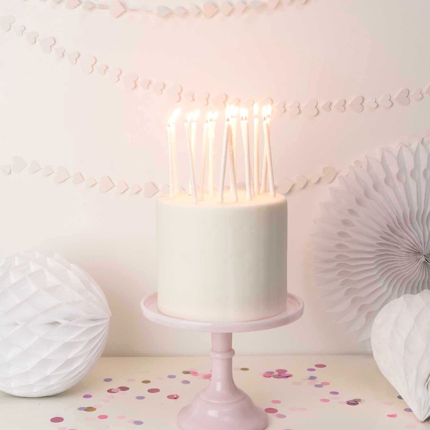 Kerzen lang 12cm, 16 Stück von Rico Design auf cremiger Torte mit brennenden Stabkerzen, pastellrosa Tortenständer und dezentem Party-Konfetti