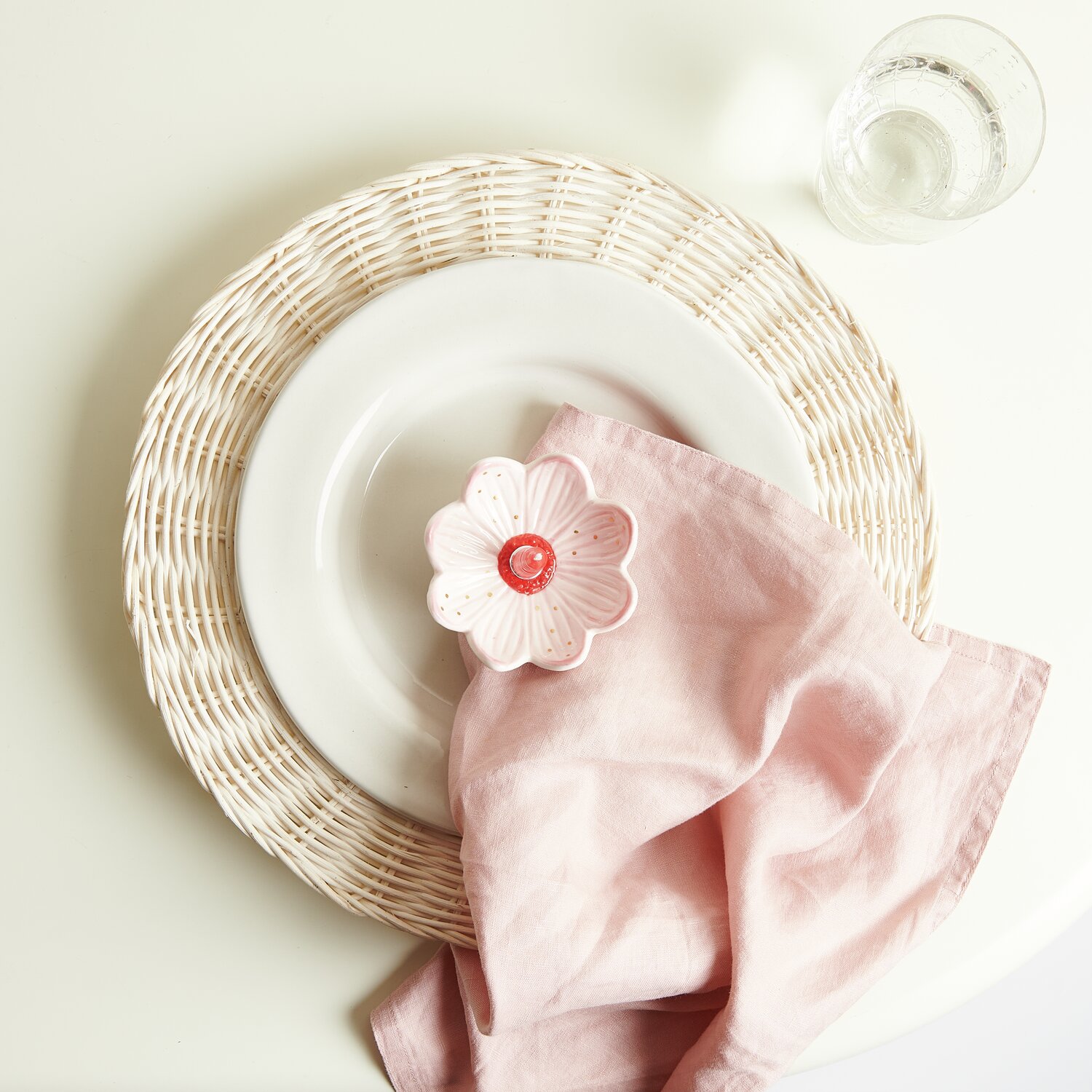 Rattan Deko-Teller Weiß mit Keramik Kerzenhalter in Blütenform, rosa Stoffserviette und Glas.