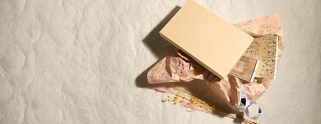 Geöffnete Geschenkbox mit dekorativem Seidenpapier, Papierprodukten und Trockenblumen auf hellem Hintergrund.