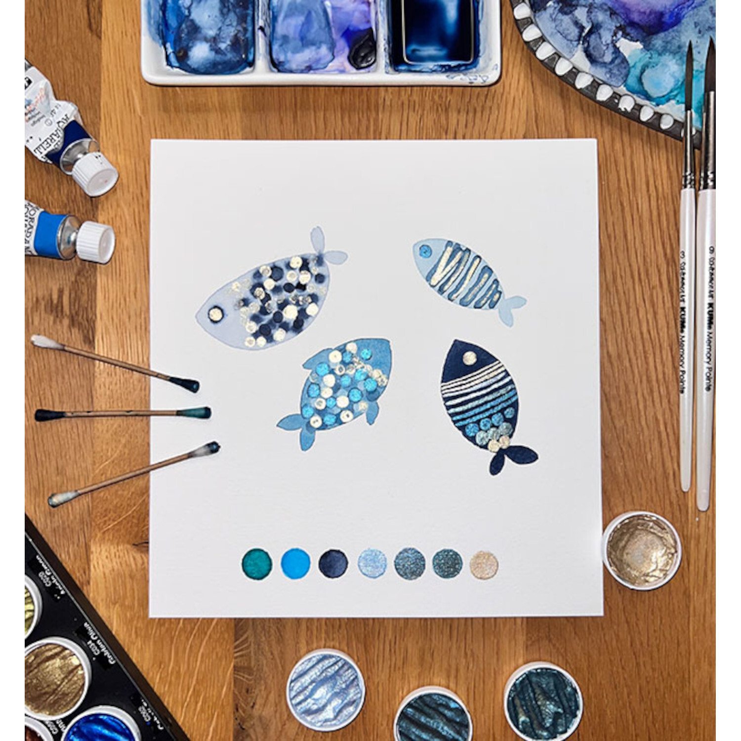 Watercolor your Mind: Kreatives Fisch-Aquarell auf weißem Papier, umgeben von blauen und silbernen Farbtönen, Pinseln und Farbpaletten.