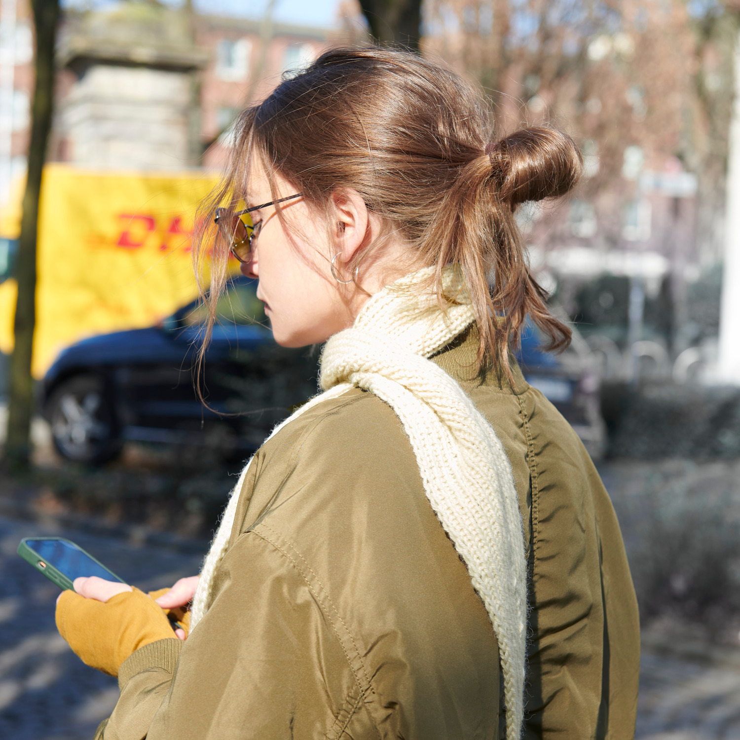 Strickset Mini Tuch Modell 04a: Frau im Freien mit beigem Schal und olivgrüner Jacke, hält ein Smartphone.