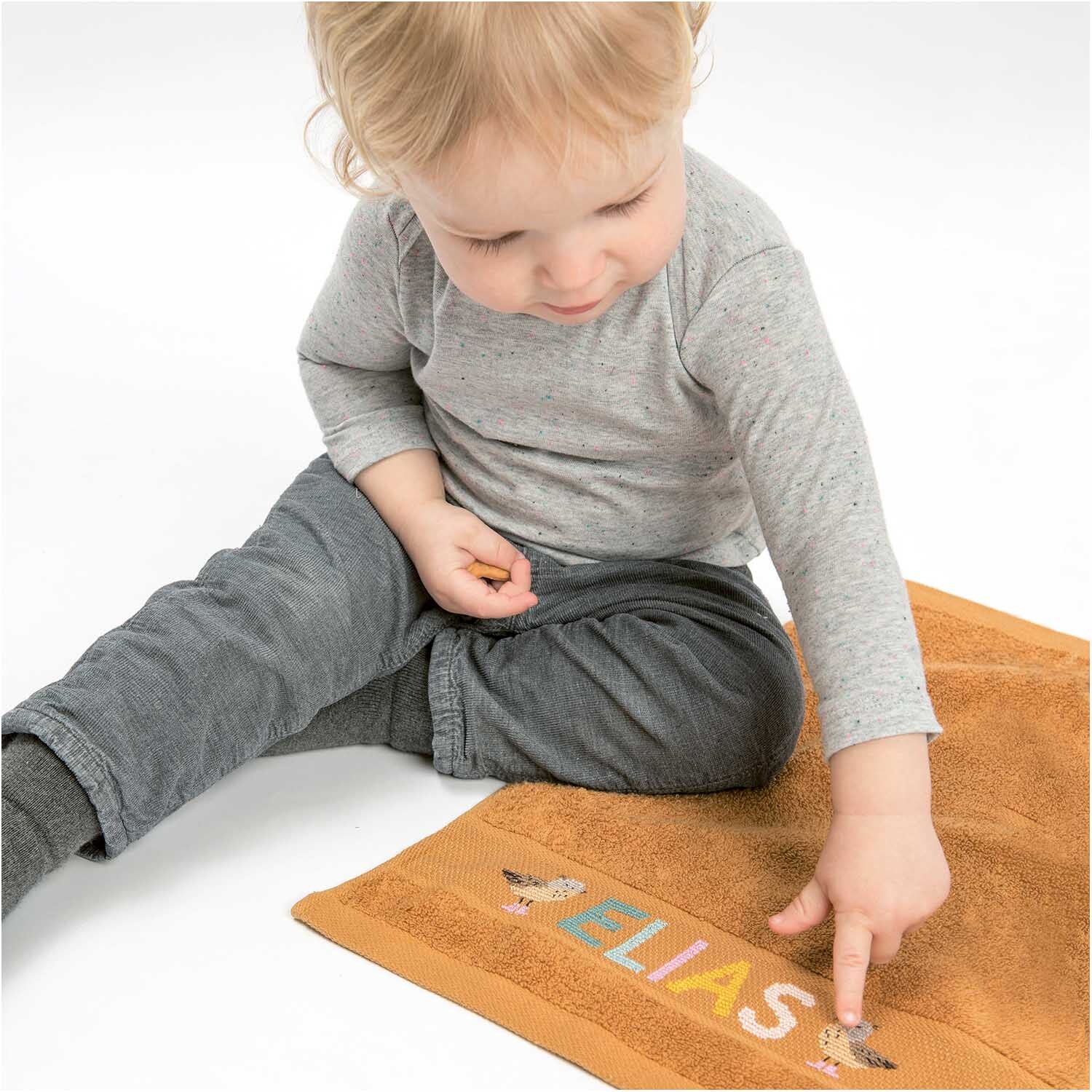 Gästehandtuch 30x50cm: Kind zeigt auf ein senf-dunkles Handtuch von Rico Design mit buntem Schriftzug.