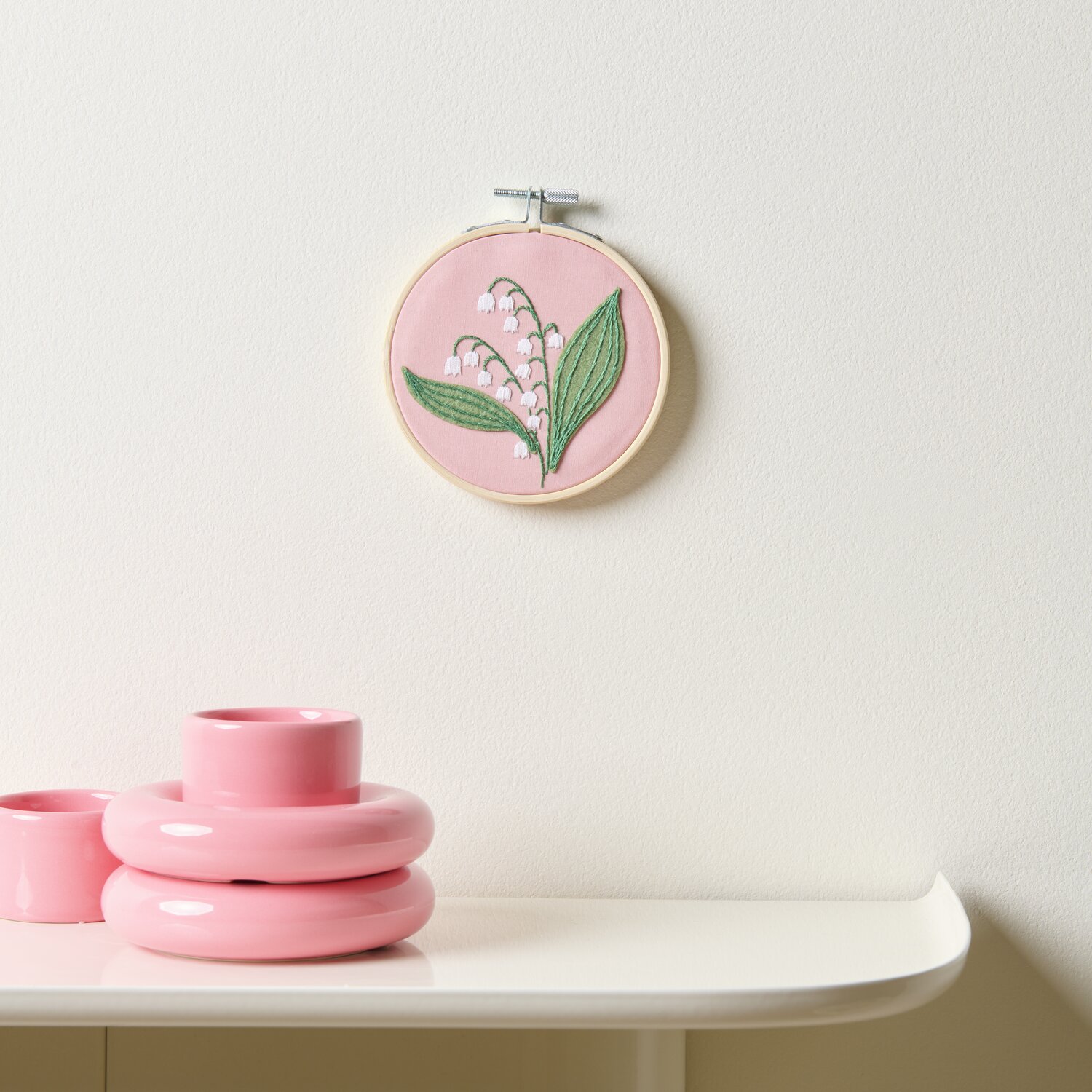 Stickpackung Vorgezeichnet Maiglöckchen auf rosa Hintergrund, Ø10cm, an weißer Wand hängend. Daneben rosa Deko-Objekte auf einem Tisch.