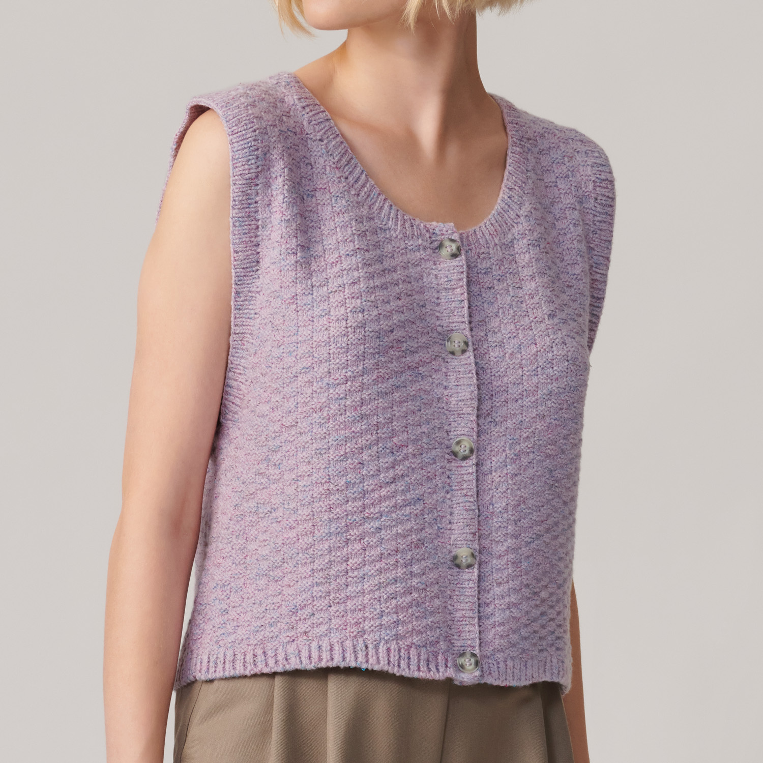 Strickanleitung Weste aus Fashion Cottony: Lila Weste mit Knöpfen, ärmellos, über beigefarbener Hose getragen.