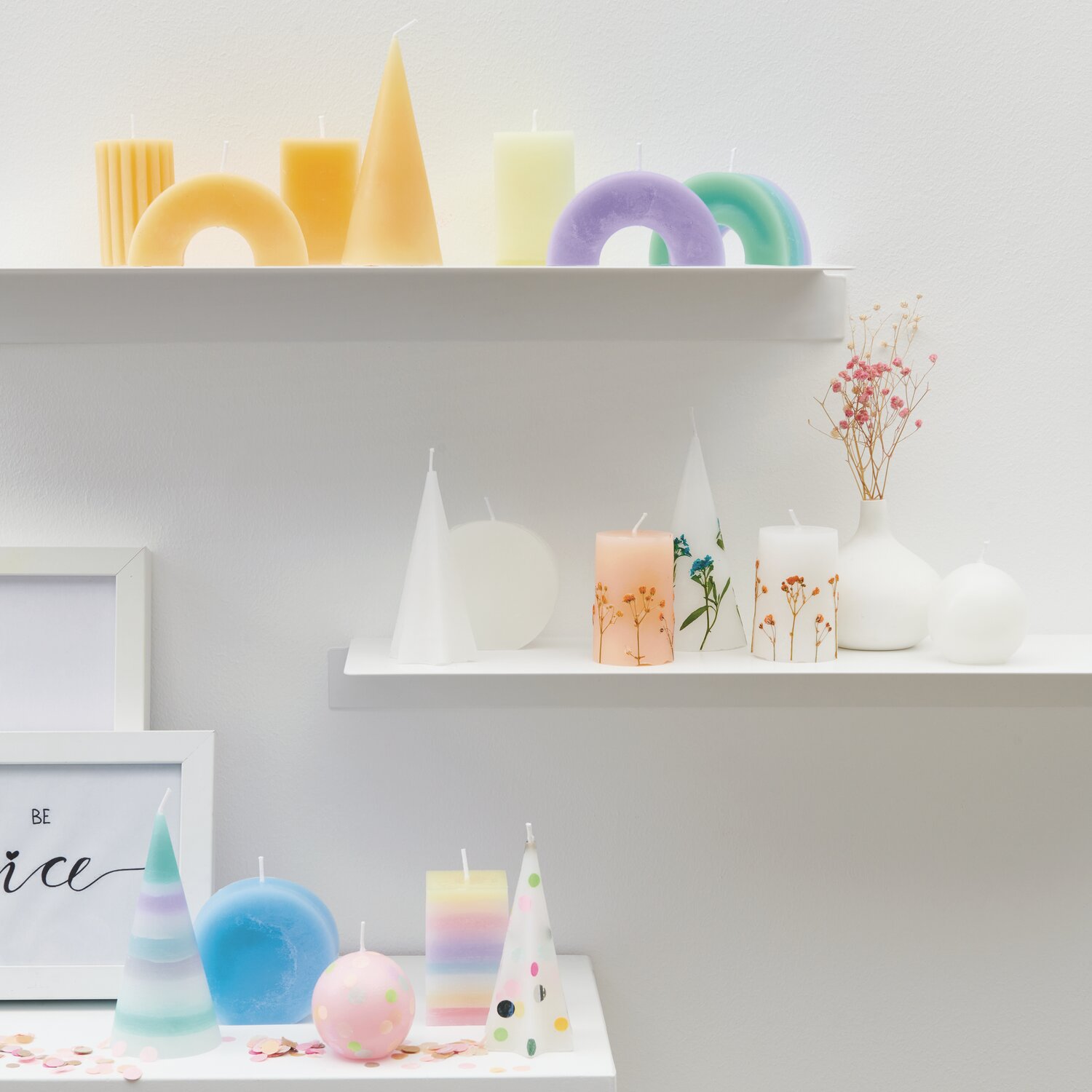Kerzenwachs Granulat Größe 300g von Rico Design Kerzenwachs Granulat: Bunte, geometrische Kerzen auf weißen Regalen, dekoriert mit Trockenblumen.