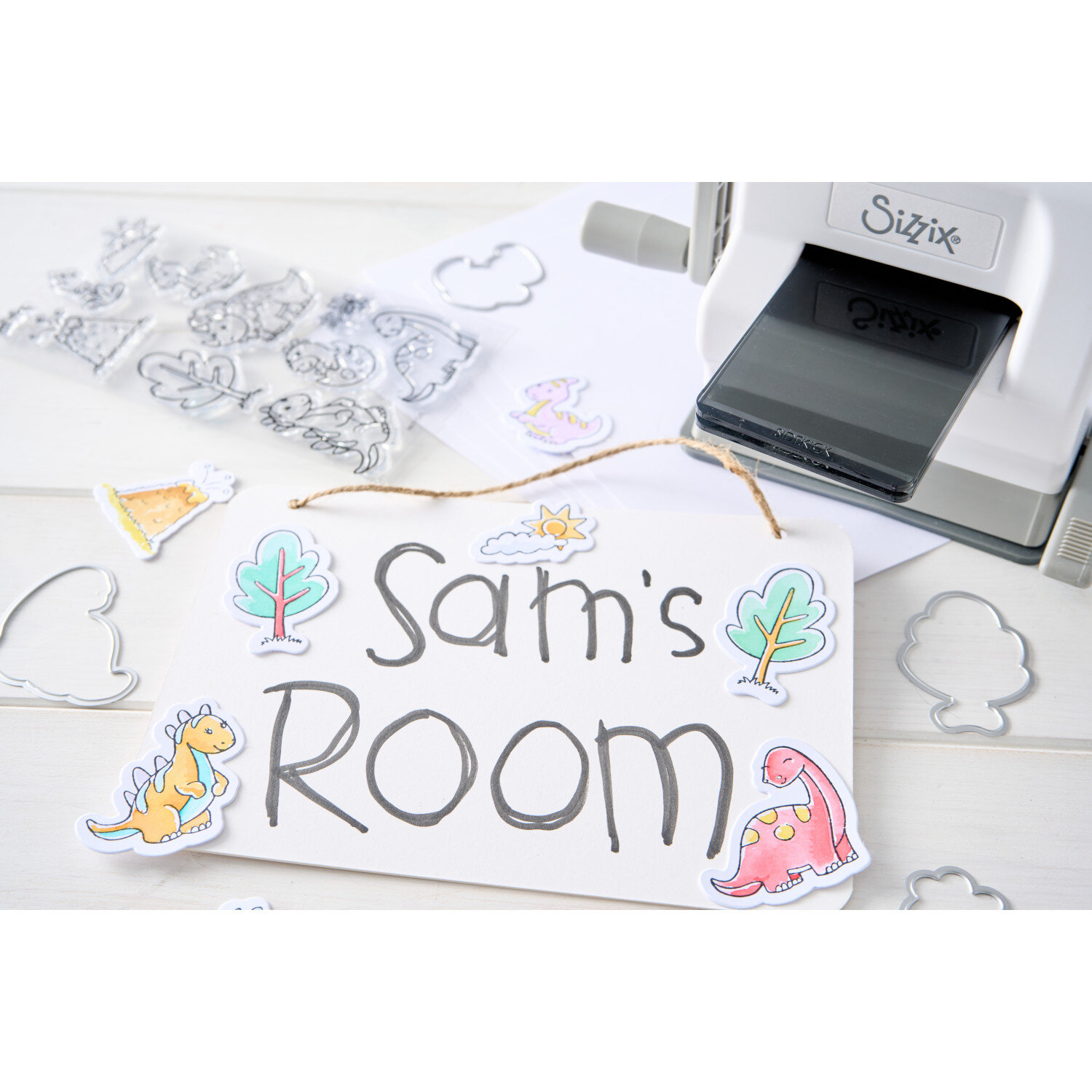 Sizzix Clear Stamps/Framelits Dinosaur Adventures Set 11 Stk zeigt ausgestanzte, bunt bemalte Dinosaurier- und Baum-Motive neben Stanze und Stempelbögen.