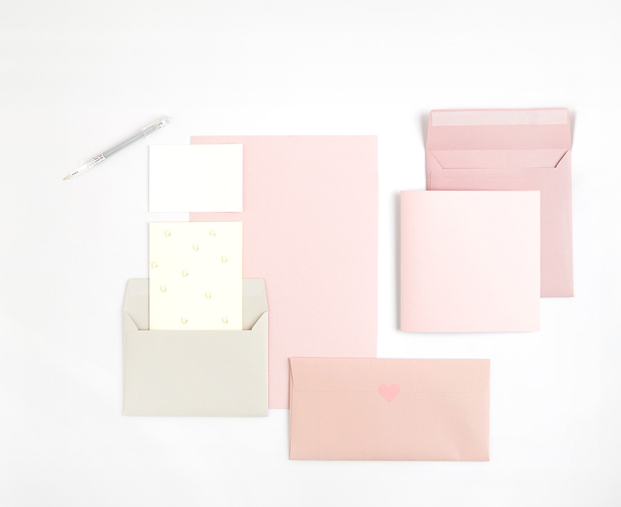 Rosa Papier darauf grauer Umschlag mit weißen Karten, sowie zwei rosa Umschläge mit heller Karte. Flatlay von Produkten.