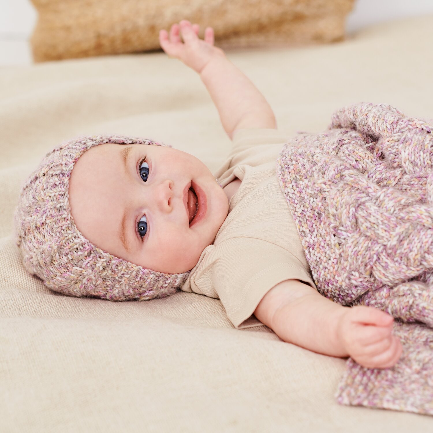 Strickset Mütze Modell 09b aus Baby 044, weich gestrickte Babymütze und passender Decke in melierter Pastelloptik, getragen von lächelndem Säugling liegend auf Decke