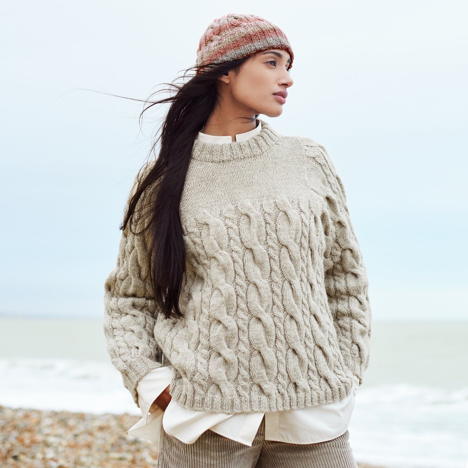 Strickset Pullover Modell 22 aus Lovewool No. 21 von Rico Design Strickset Pullover Modell 22 aus Lovewool No. 21, beige, am Strand getragen, mit Zopfmuster.