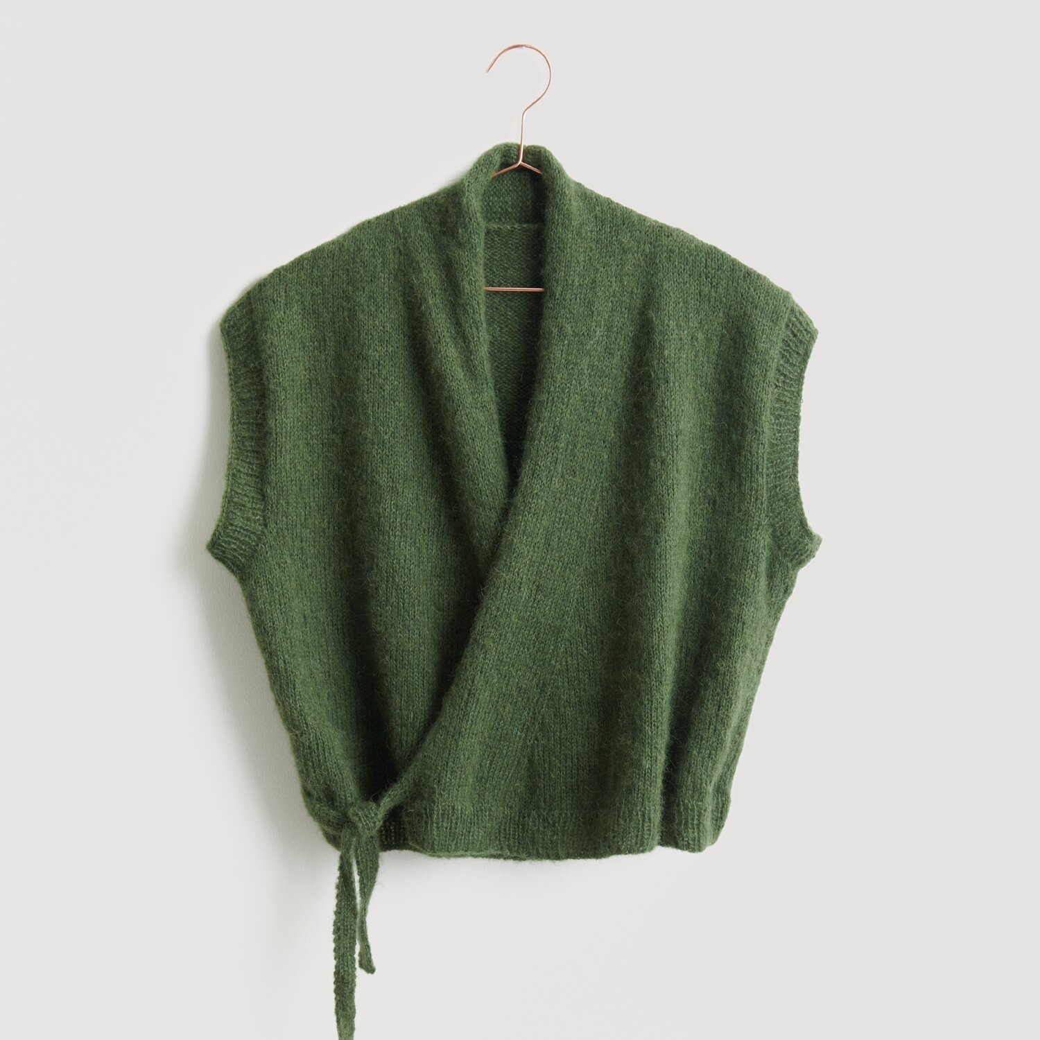 Lovewool No. 21 Herbst-Winter Sprachauswahl deutsch von Rico Design Grüne Strickweste von Modell Legeware, präsentiert auf einem Kleiderbügel. Die Weste hat kurze Ärmel und einen Wickelstil mit Bindeband, ideal für einen lässigen Look.