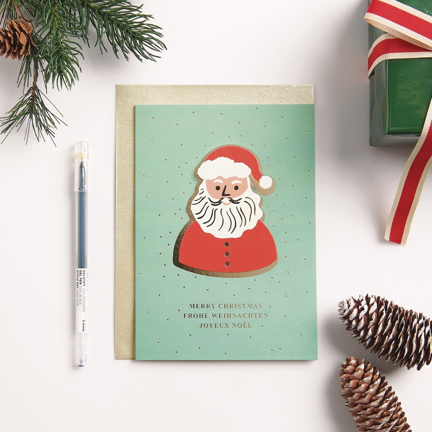 3D-Kartenset Weihnachtsmann B6: Weihnachtskarte mit rotem Santa-Motiv auf grünem Hintergrund, umgeben von Tannenzweigen und Geschenk.