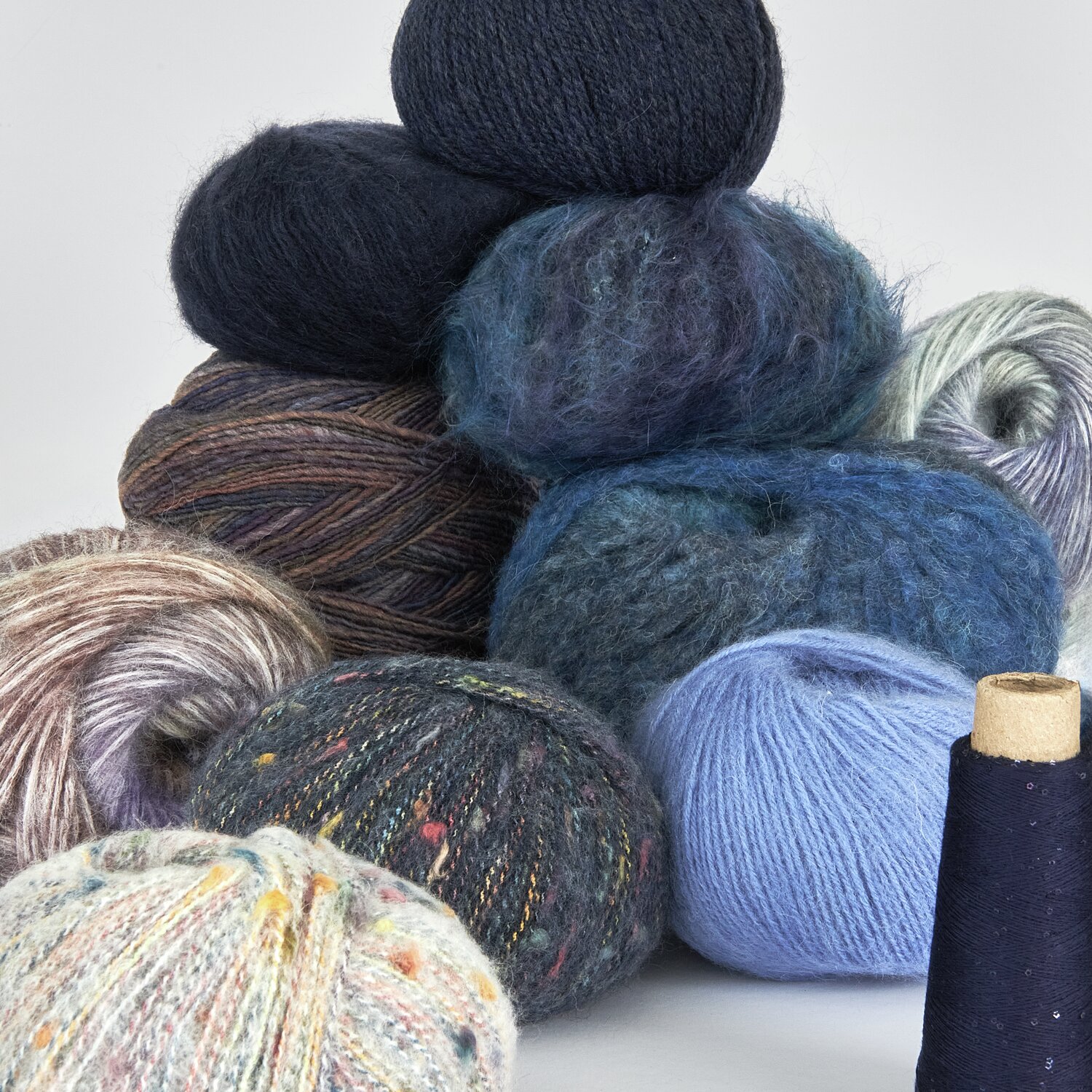 Lovewool No. 21 Herbst-Winter Sprachauswahl deutsch von Rico Design Ein Stapel aus verschiedenen Wollknäueln in Blau-, Grau- und Brauntönen, darunter flauschige und melierte Texturen. Ideal für Strickprojekte. Marke: Unbekannt.