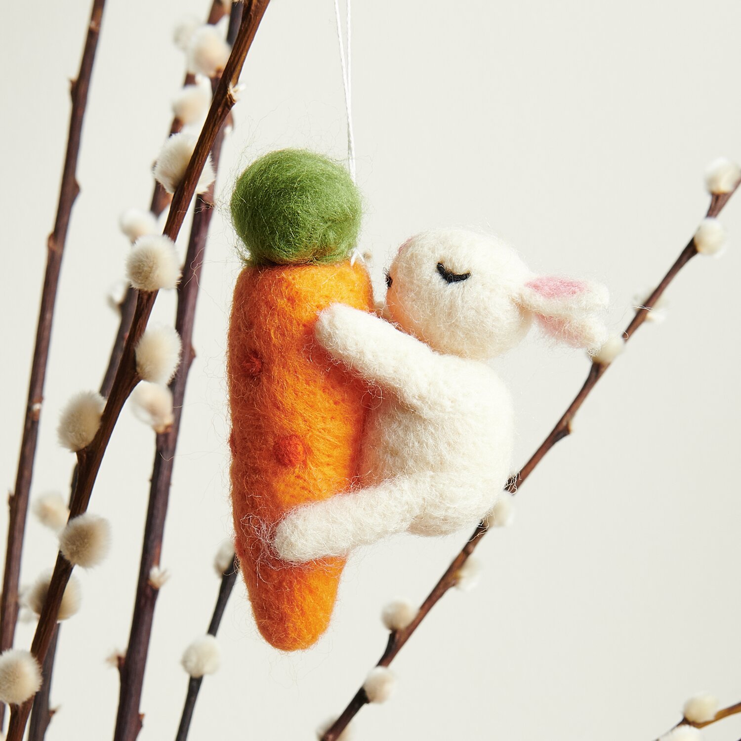 Aufhänger Hase auf Möhre: Niedlicher Filzhase klammert sich an orangefarbene Karotte, dekorativ an Zweigen hängend. Größe: 10,7x3,8x7,8cm.