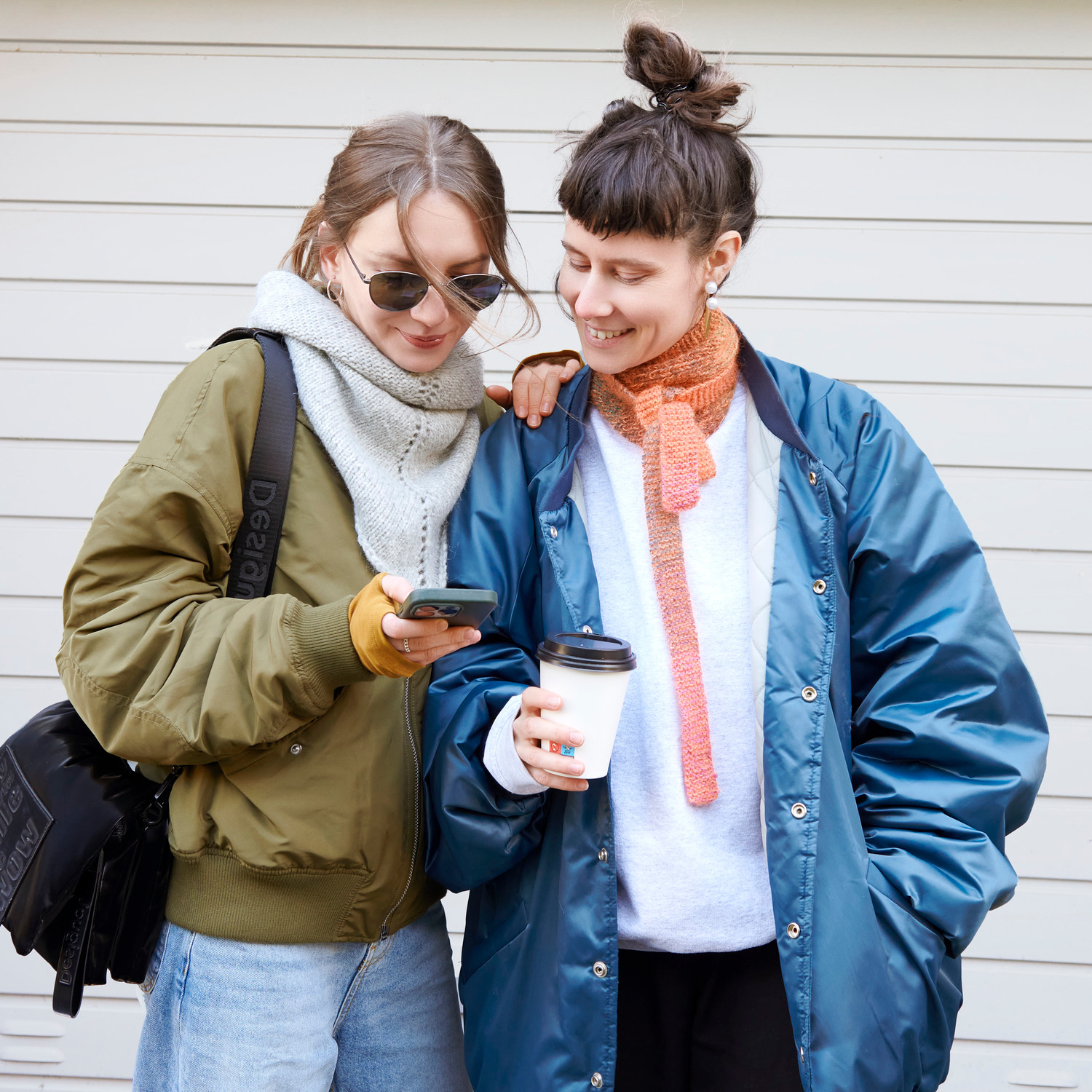 Strickset Dreieckstuch Modell 05a: Zwei Frauen in Jacken, eine mit grauem, die andere mit orangefarbenem Tuch, lächeln und schauen auf ein Handy.