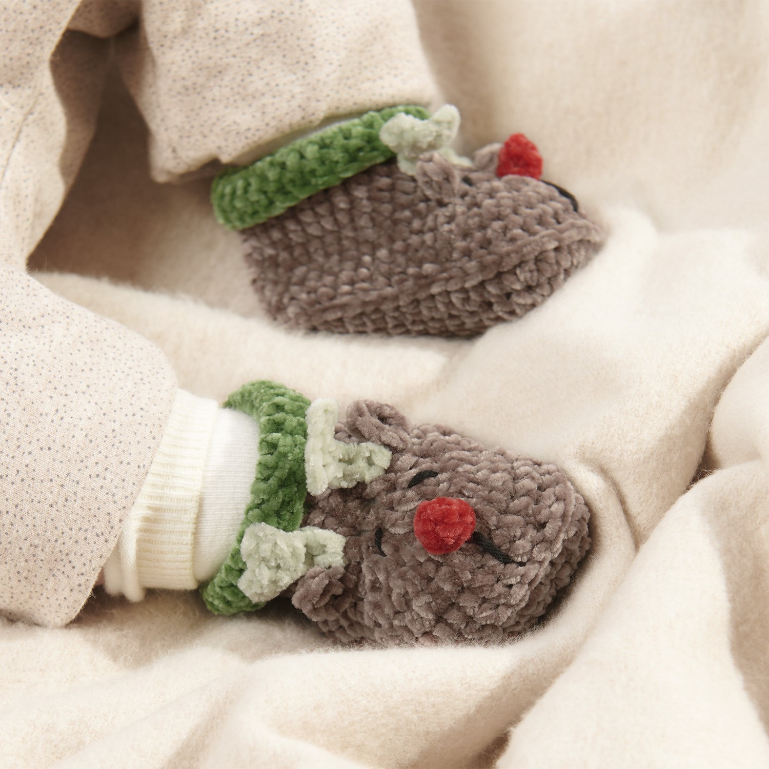 Häkelanleitung Baby Booties Rentier: Niedliche braune Rentier-Babyschuhe mit grünen Details und roter Nase auf beigem Stoff. Perfekt für die Weihnachtszeit.