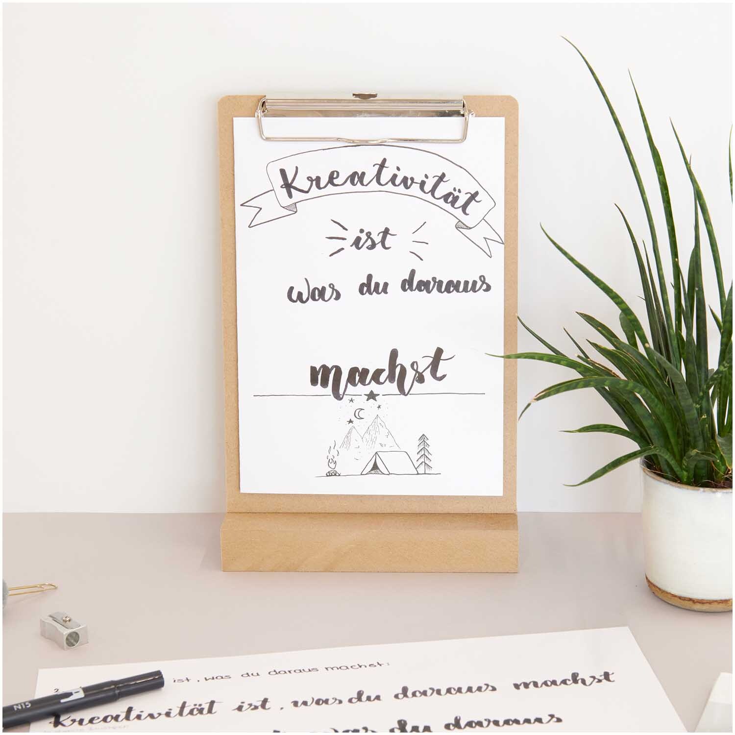 Klemmbrett Natur DIN A4 mit kreativem Schriftzug, umgeben von Pflanzen auf einem Schreibtisch.