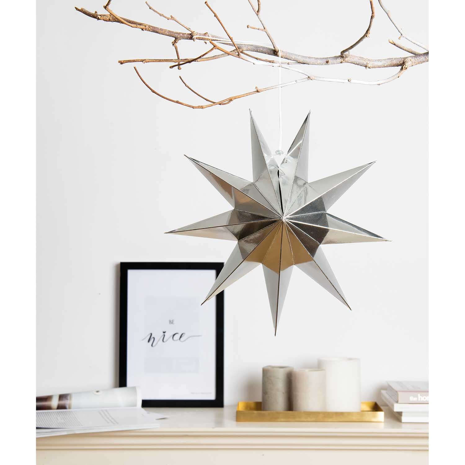 Papierstern auffaltbar 28cm, silber glänzend, hängt an einem Ast über einem Regal mit Kerzen und Kunstwerk. Ideal für festliche Dekorationen.