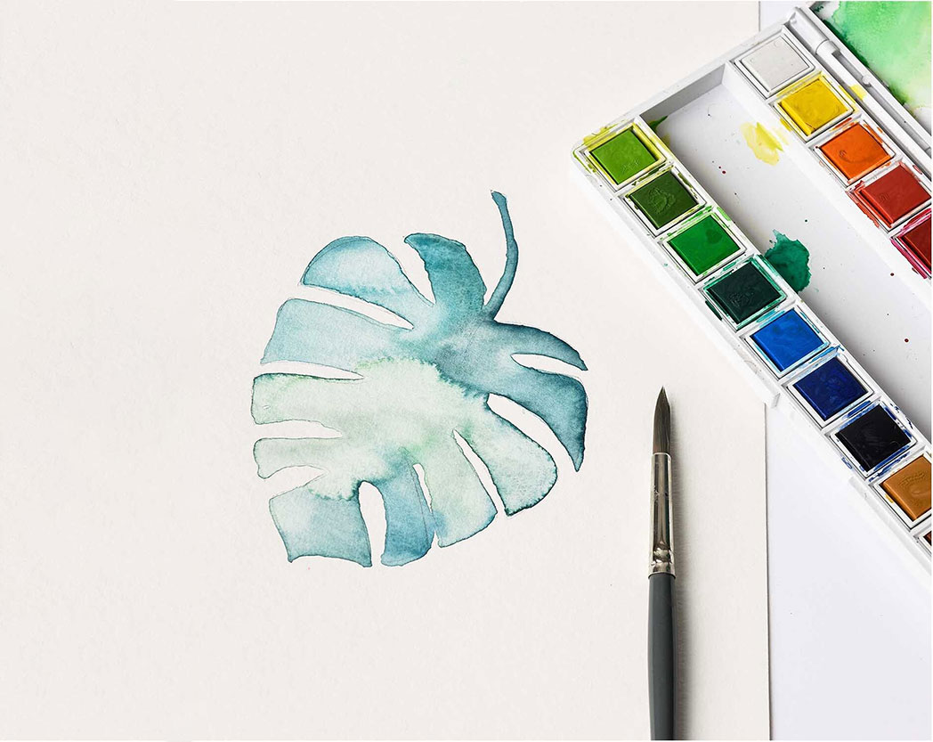 Ein Monsterablatt mit grün und blauer Aquarellfarbe gemalt, daneben ein Pinsel und ein Aquarellfarbkasten. Flatlay von Produkten.