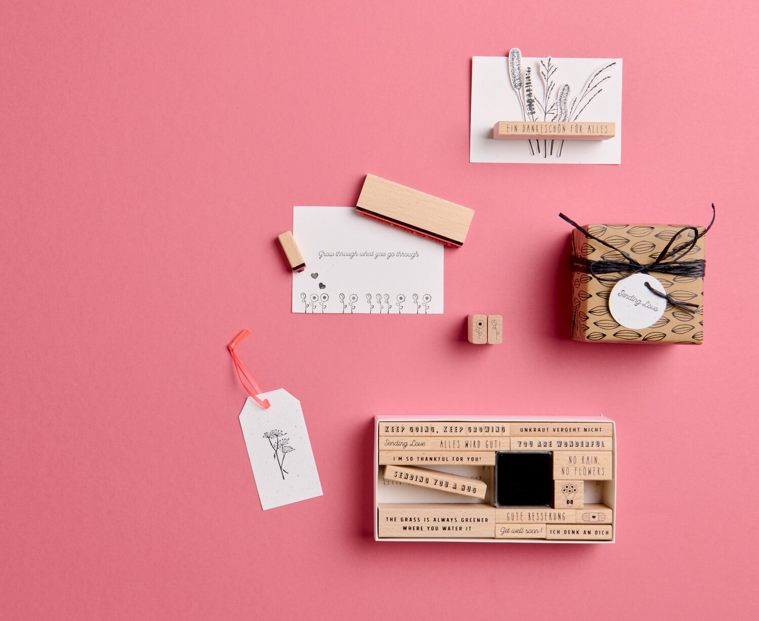 Stempelset Motivation 20-teilig zeigt Holzstempel von Rico Design mit Sprüchen, Zahlen und Symbolen, arrangiert auf rosa Untergrund mit Karten und Geschenkbox